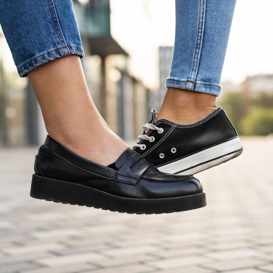 A person wearing black leather platform penny loafers with white laces and white midsoles, paired with blue jeans, photographed outdoors on a blurred paved walkway.