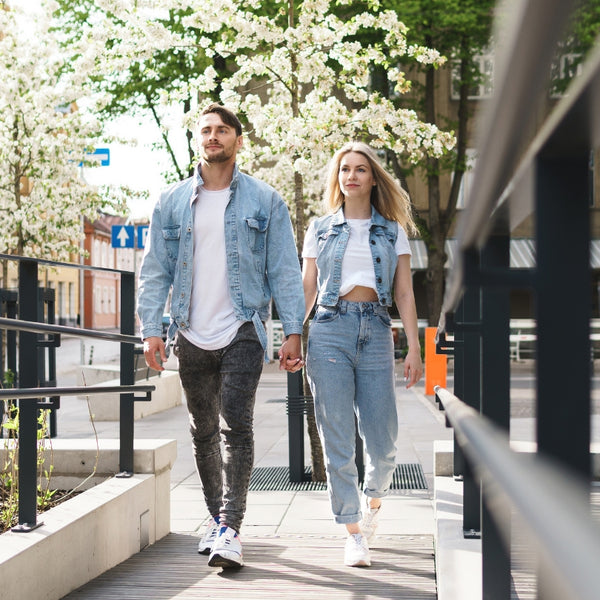 A man and a woman walking hand-in-hand on an outdoor urban pathway lined with blooming white trees. The man wears a light blue denim jacket over a white t-shirt and dark gray jeans, while the woman wears a matching light blue cropped denim jacket over a white crop top and light blue jeans.