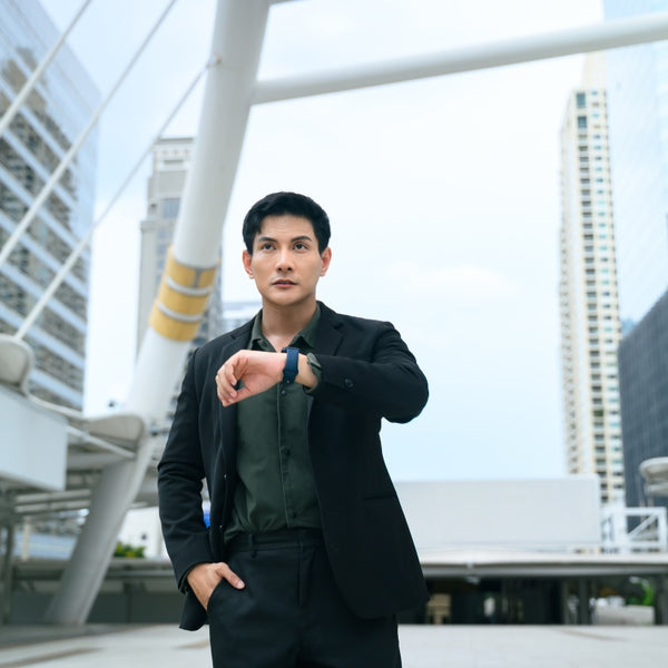 A man in a black blazer and dark green shirt checks his wristwatch outdoors in a modern city setting with white architectural structures and skyscrapers in the background.