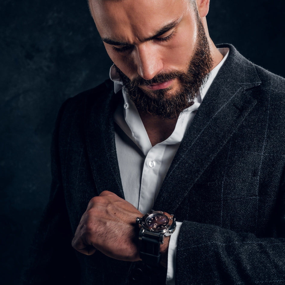 A close-up lifestyle shot of a bearded man in a dark gray blazer and white shirt looking down at a rugged black wristwatch with a brown dial, set against a dark textured background.