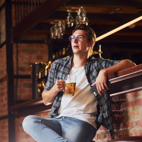 A man wearing round metal-framed eyeglasses sits in a dimly lit bar or brewery setting, holding a glass of beer. He is dressed in a white t-shirt, a dark gray plaid flannel overshirt, and blue jeans.