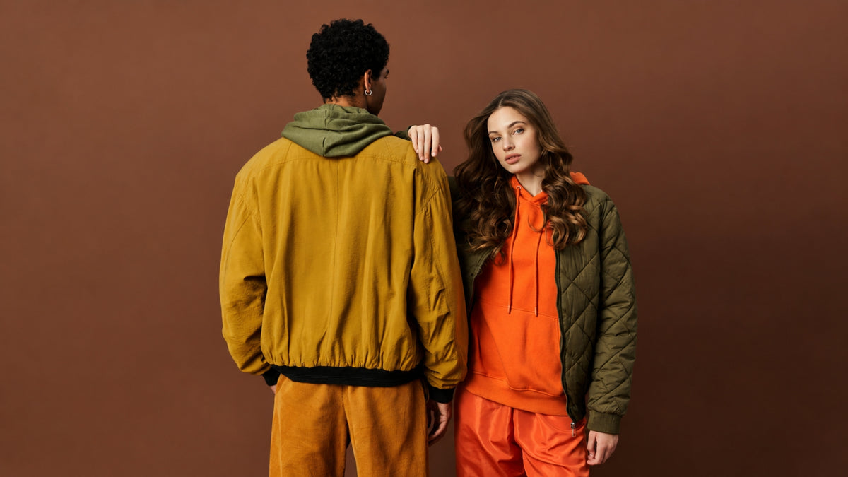 Two models standing against a solid brown studio backdrop. The model on the left is shown from behind wearing a mustard yellow jacket with an olive green hood and matching yellow pants.
