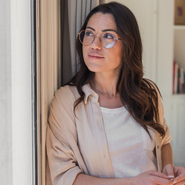 A woman with long brown hair and round metal-framed eyeglasses, wearing a beige button-up shirt over a white top, leaning against a window in a bright indoor setting.