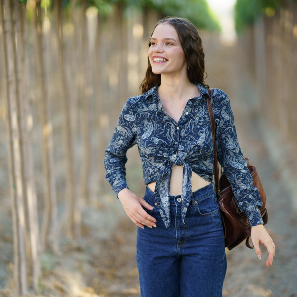 A woman standing in a vineyard wearing a blue and white patterned long-sleeve crop top tied at the waist, blue jeans, and carrying a brown leather shoulder bag.