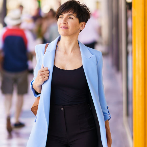 A woman walking outdoors wearing a light blue blazer over a black top and black pants, with a brown shoulder bag.