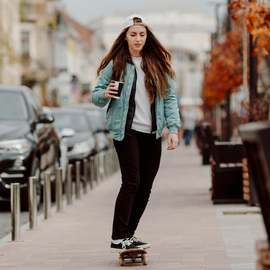 A woman wearing a light blue jacket, black pants, and a white cap stands on a skateboard on a city sidewalk, holding a coffee cup.