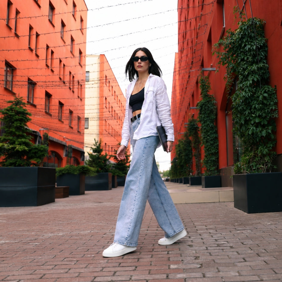 A woman walking outdoors on a brick pathway between tall orange-red buildings, wearing a white button-up shirt, light-wash wide-leg jeans, a black top, and white sneakers.